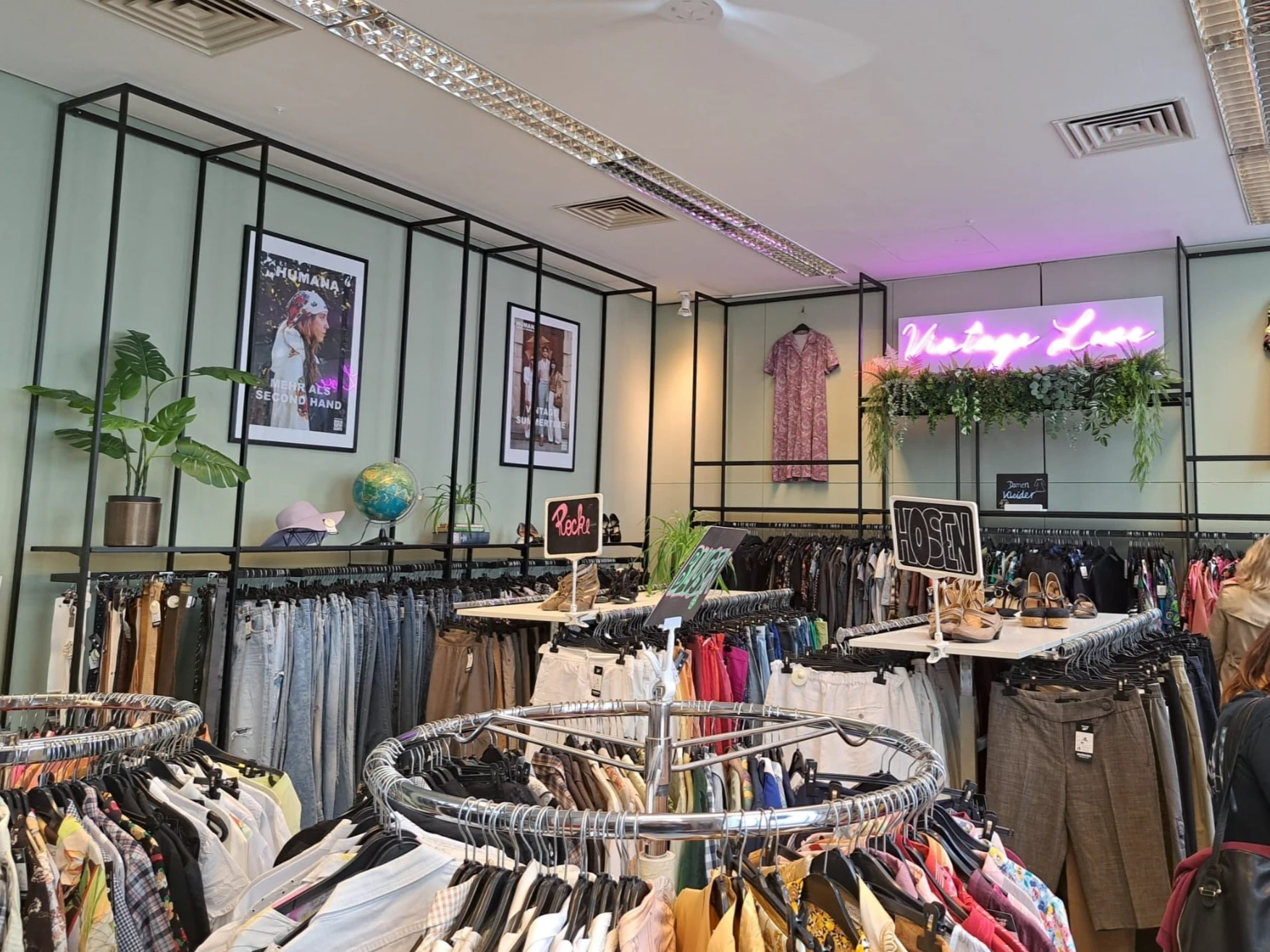 Blick in einen hellen Secondhand-Laden mit Kleiderständern, runden Kleiderständern im Vordergrund und einem 'Vintage Love' Neonschild.